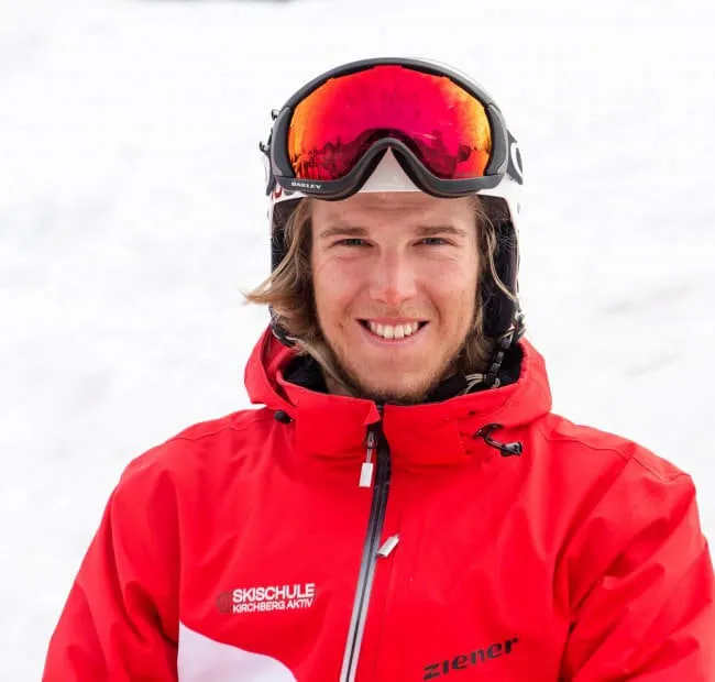 Lächelnder Skifahrer in roter Jacke mit Sonnenbrille im Schnee.